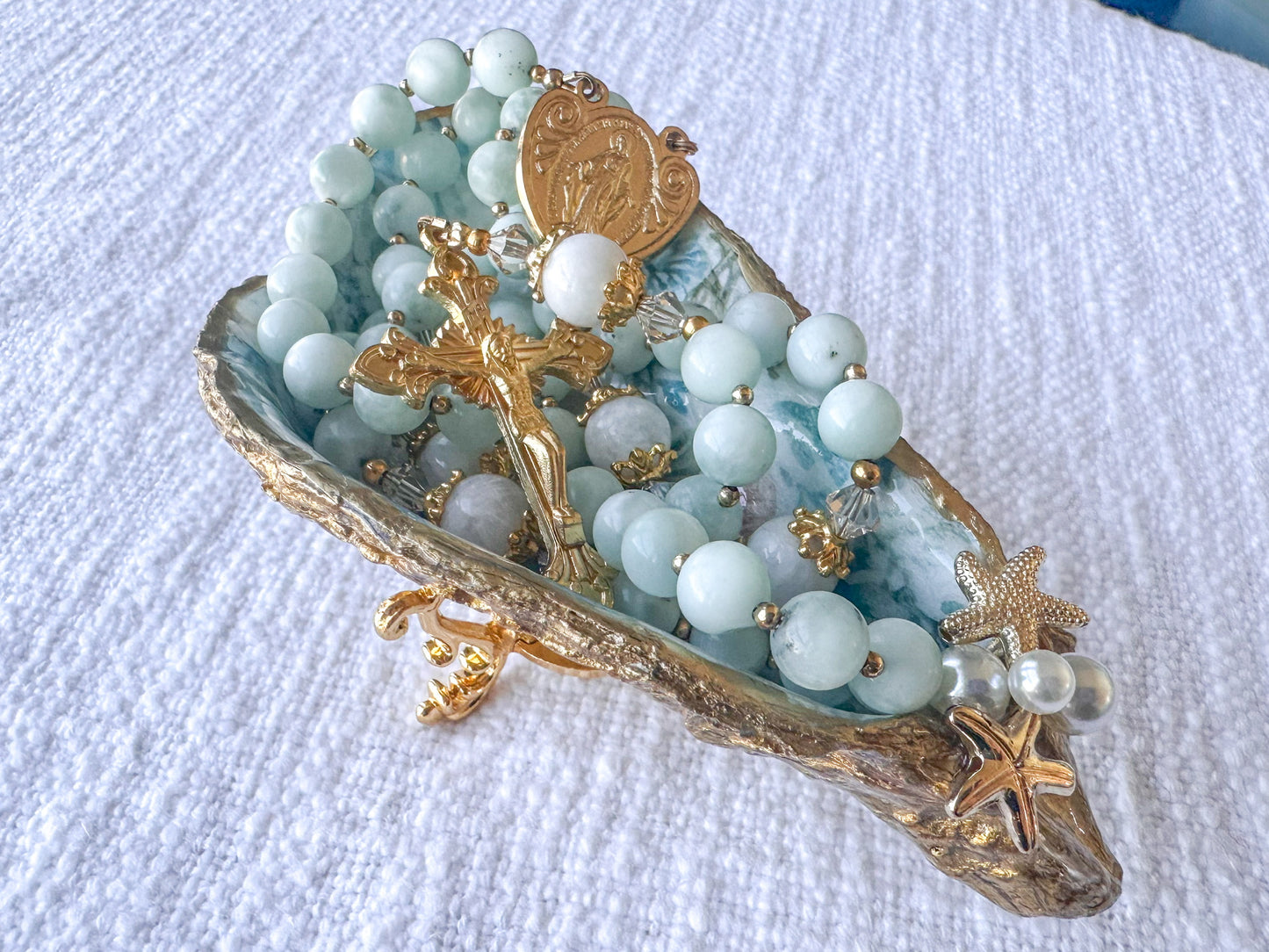 Decorative dish with rosary beads, gold cross, and starfish on a white textured surface