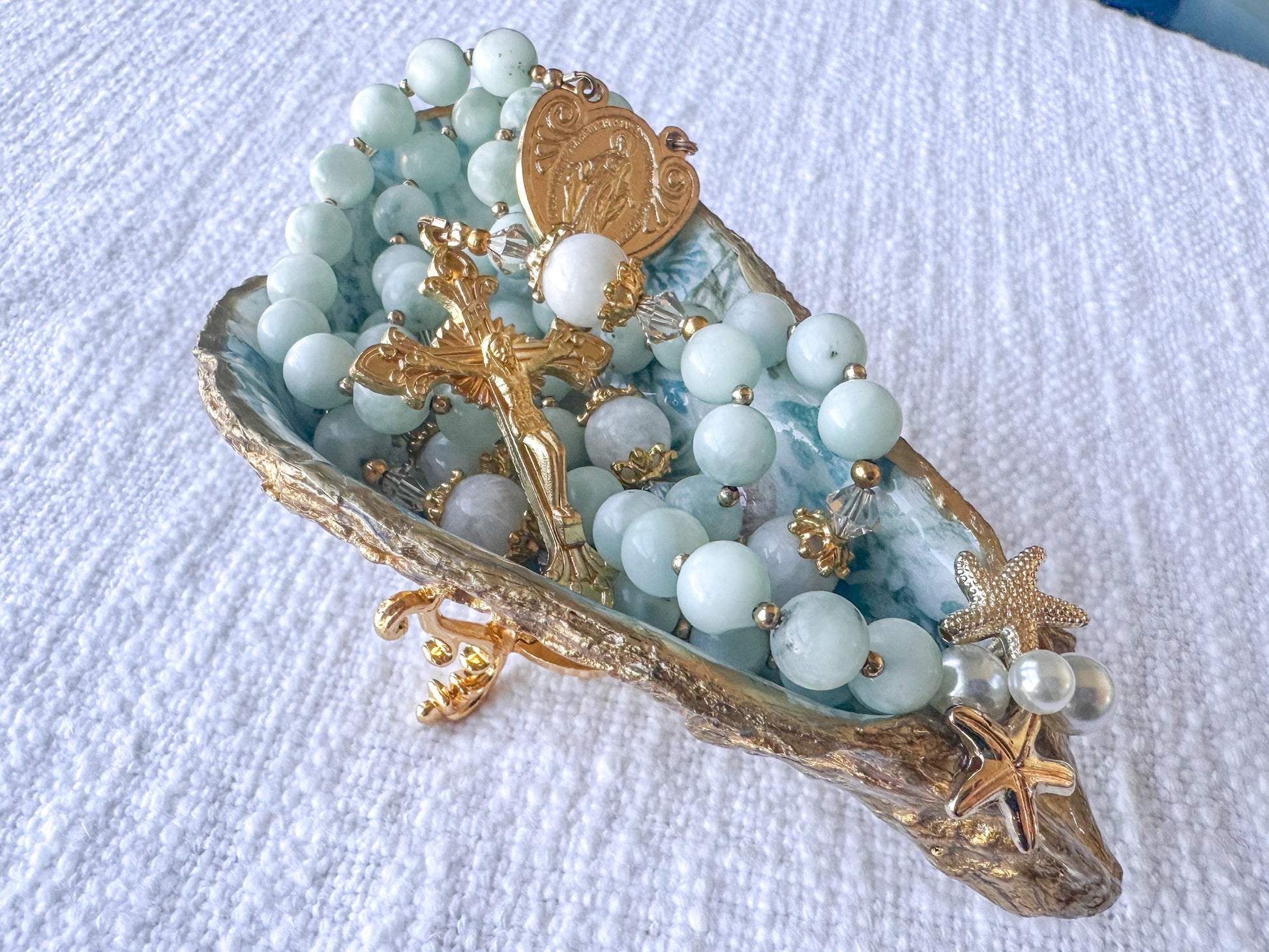 Decorative dish with rosary beads, gold cross, and starfish on a white textured surface